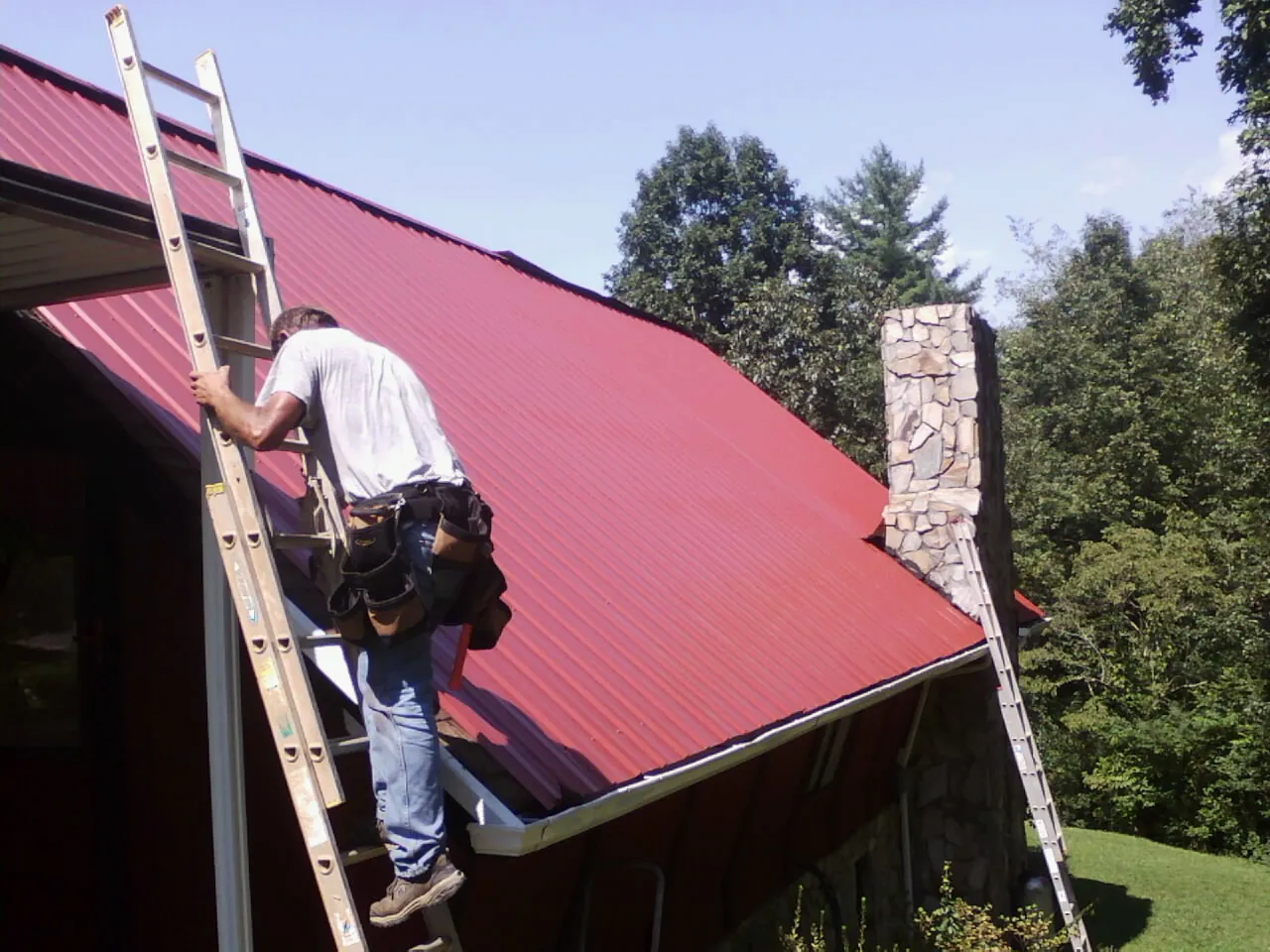 Roofer on a ladder beside a red metal roof and stone chimney — Soffit & Fascia Repair by Roofing Brothers in Twin Valley
